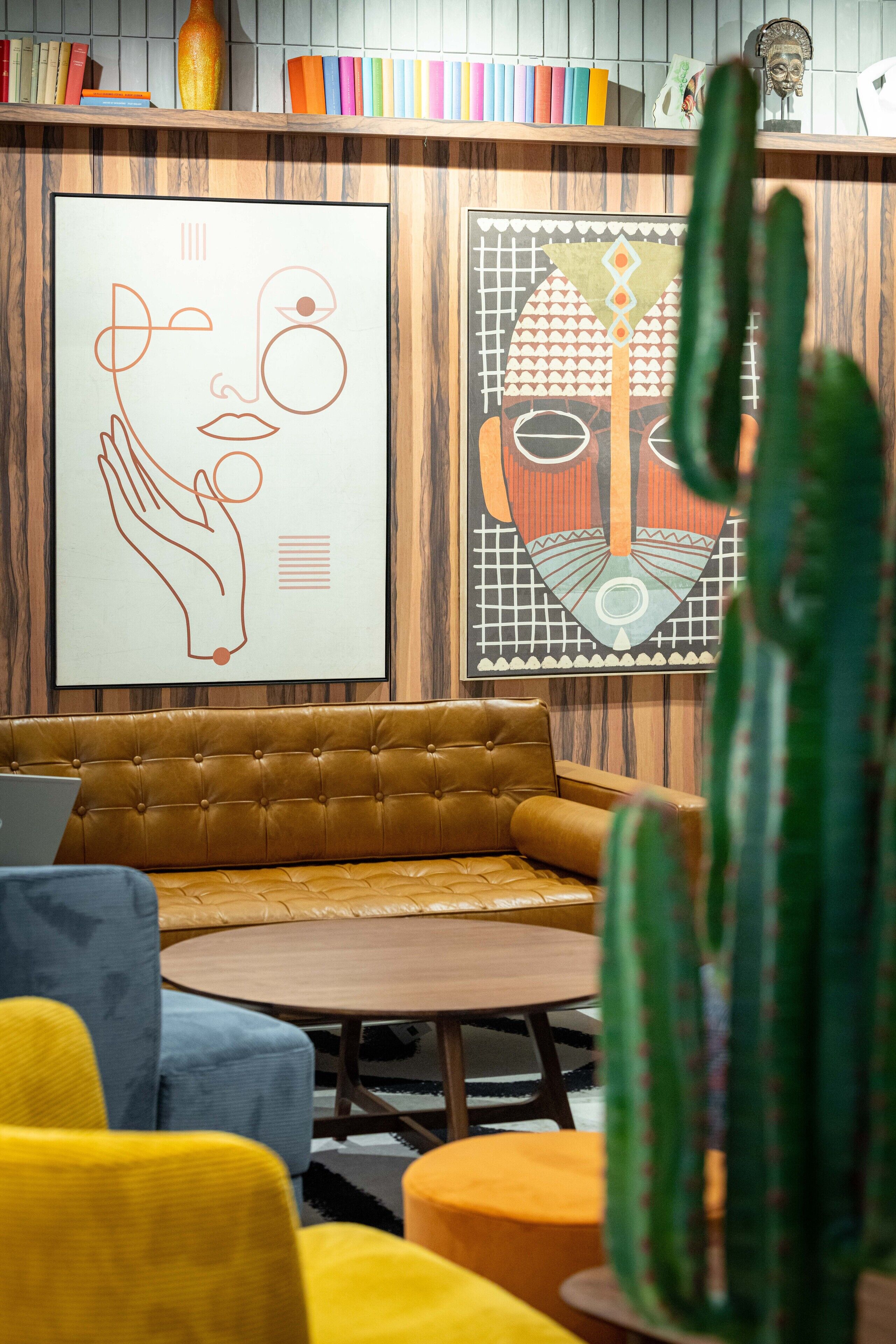 Un espace de détente moderne avec un canapé marron, une table ronde en bois et des chaises colorées. On peut voir des livres sur une étagère et un cactus au premier plan.