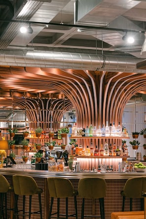 Zona de bar con techo de madera curvada, taburetes y una variedad de botellas y vasos sobre la barra.