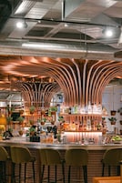 Zona de bar con techo de madera curvada, taburetes y una variedad de botellas y vasos sobre la barra.