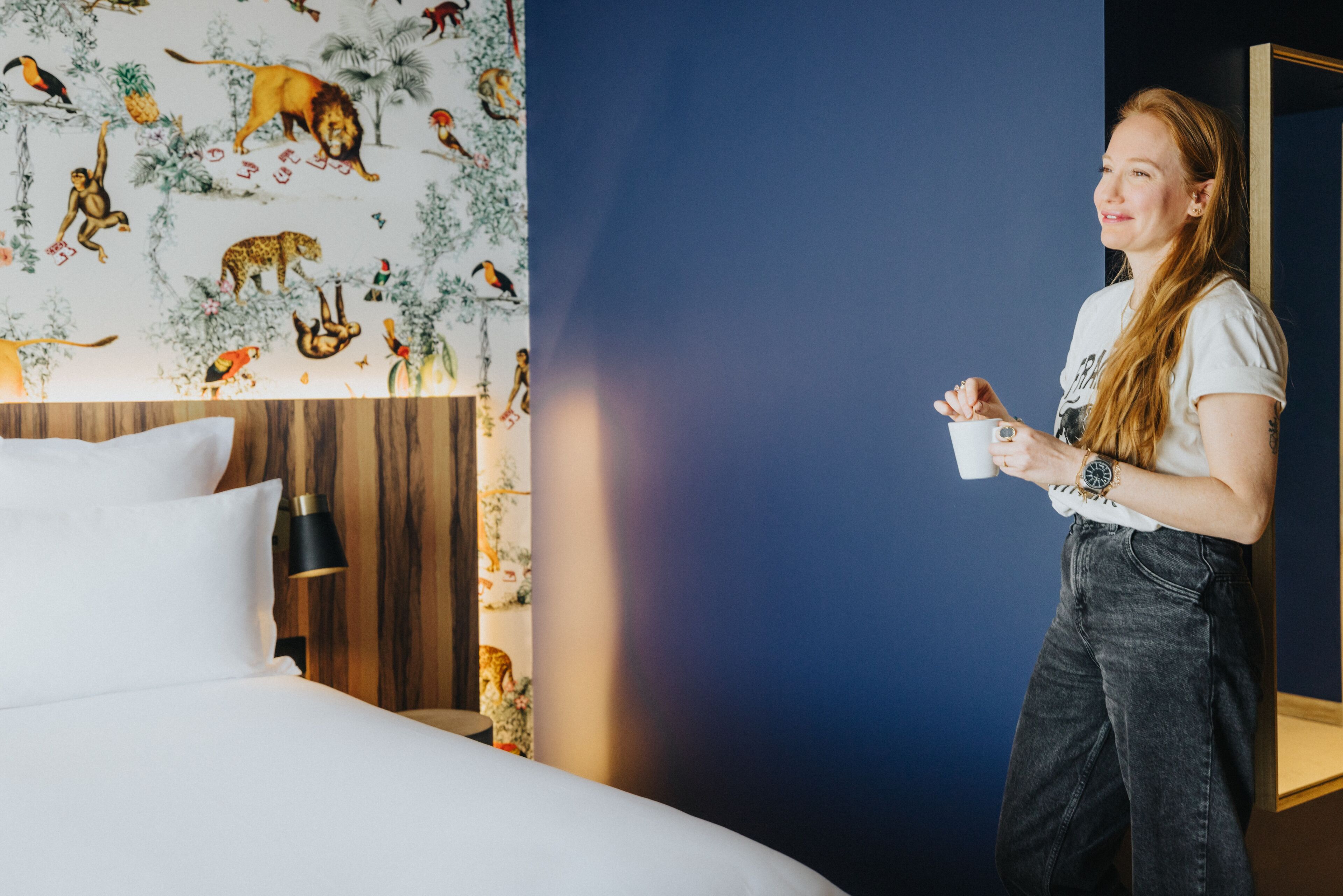 Ein Hotelzimmer mit einem Bett, einem Nachttisch, einer Lampe und einer Wand mit Dschungel-Tapete.