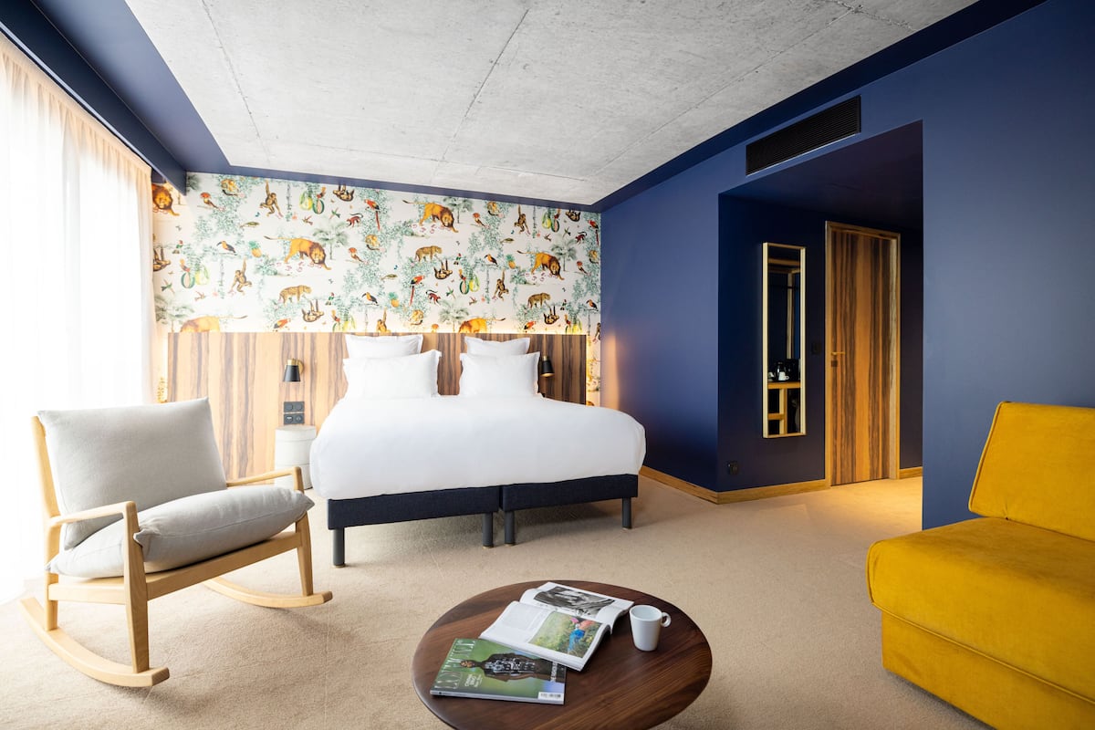 A modern hotel room with a bed, a rocking chair, a yellow sofa, and a coffee table with a magazine.