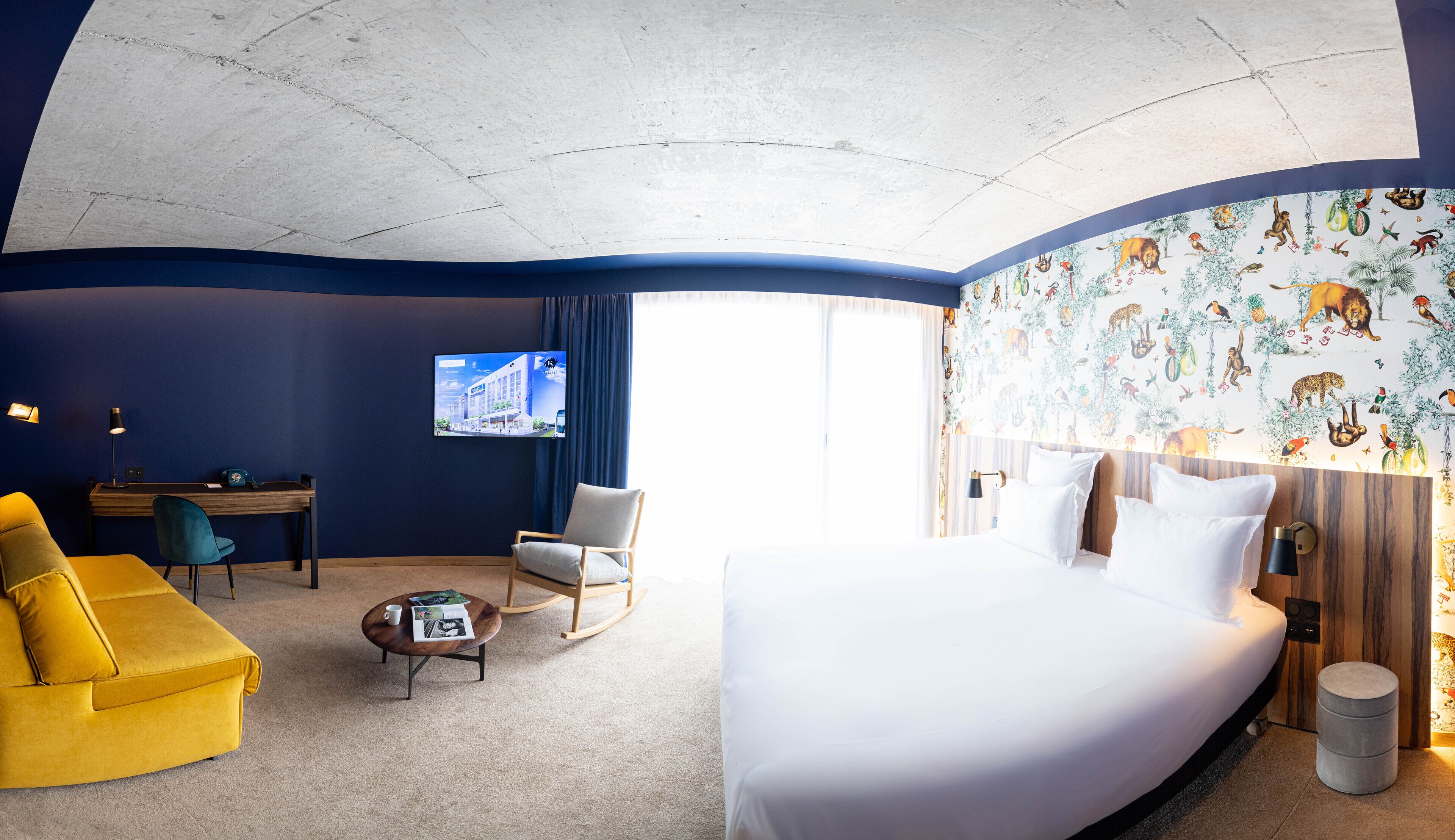 A hotel room with a bed, a desk, a chair, a sofa, a TV, and a mural on the wall.