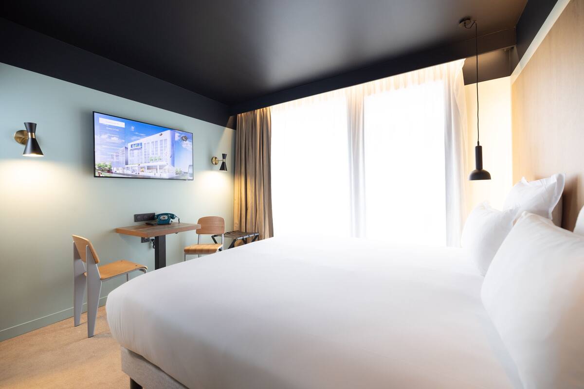 A modern hotel room with a large bed, a desk with two chairs, a flat-screen TV on the wall, and a window with curtains.