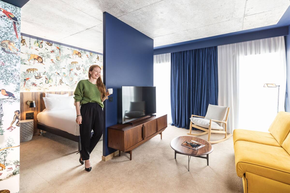 A modern hotel room with a bed, a TV, a sofa, and a chair.
