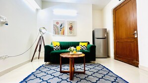 Un espacio habitable pequeño con un sofá verde, una mesita redonda, un refrigerador moderno y una puerta de madera.