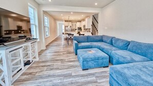 A spacious living area with a blue sectional sofa, a kitchen with white cabinets, and a staircase leading to another level. - Bayside * 5BR Luxury Home (Selbyville)
