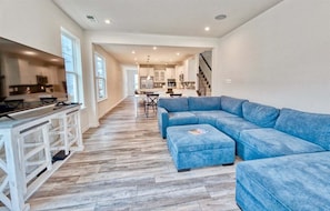 A spacious living area with a blue sectional sofa, a kitchen with white cabinets, and a staircase leading to another level.