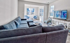 Un salon moderne avec un canapé d’angle gris, un téléviseur à écran plat et une grande fenêtre donnant sur des immeubles.
