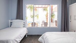 Une chambre avec deux lits, une fenêtre avec des rideaux, une armoire et une vue sur une terrasse extérieure aménagée avec des meubles et des plantes.
