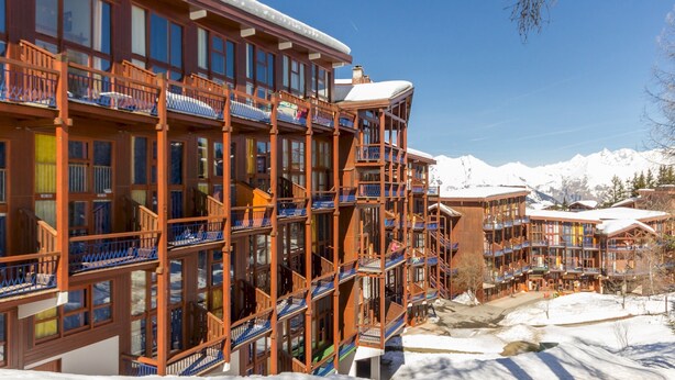 Un hôtel à plusieurs étages doté de balcons, de grandes fenêtres et surplombant un paysage montagneux enneigé.