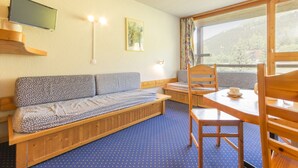 Quarto com um banco de madeira, uma mesa com cadeiras, uma TV fixada na parede e uma janela com vista para o exterior.