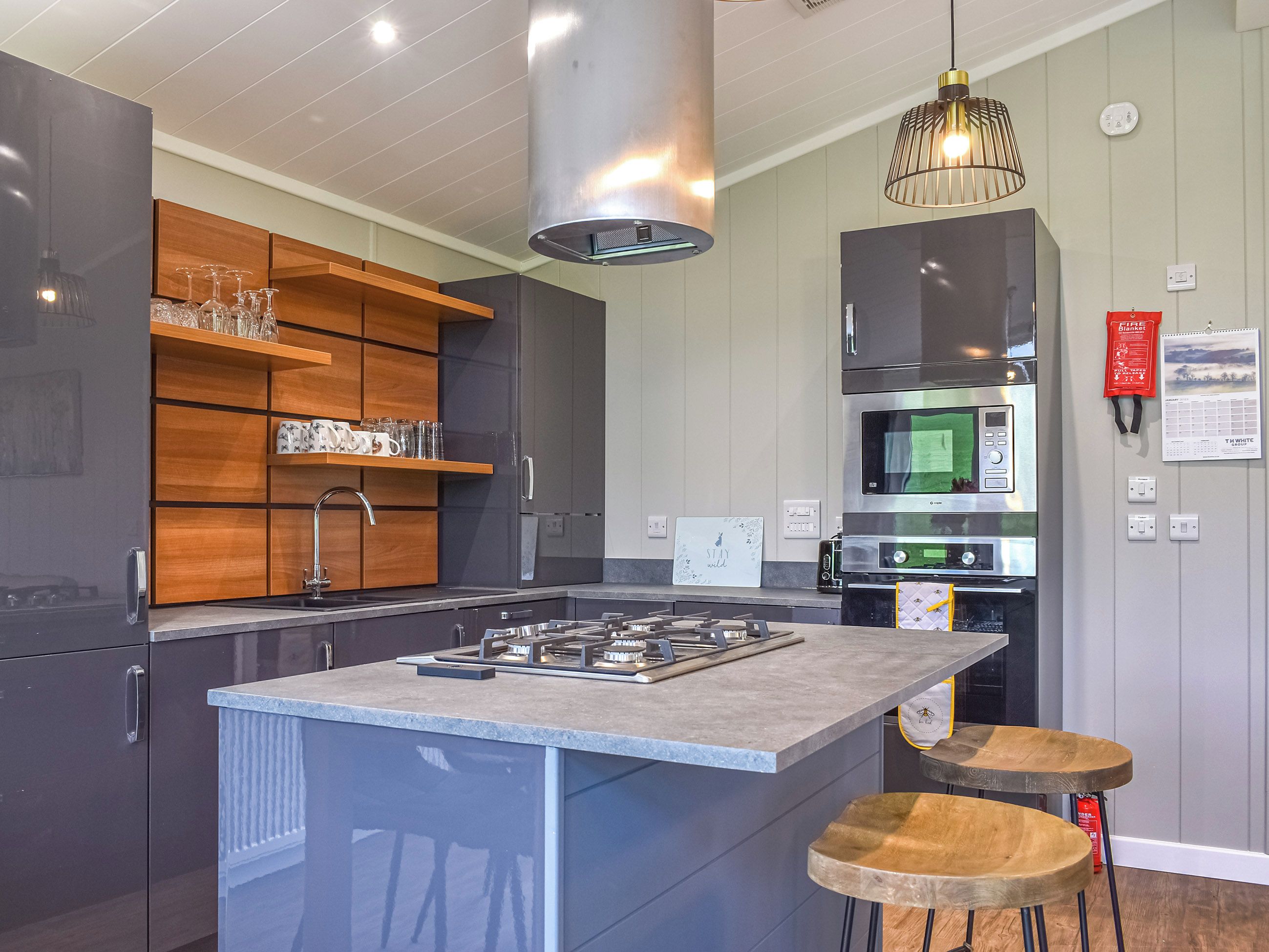 A modern kitchen with a central island, stainless steel appliances, wooden shelves, and a hanging light fixture.