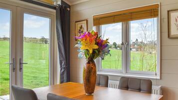 A dining area with a table, chairs, a vase with flowers, and a view of a green field through large windows.