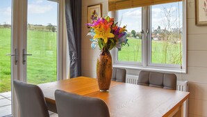A dining area with a table, chairs, a vase with flowers, and a view of a green field through large windows. - Whitey Top Country Lodge (Pentridge)