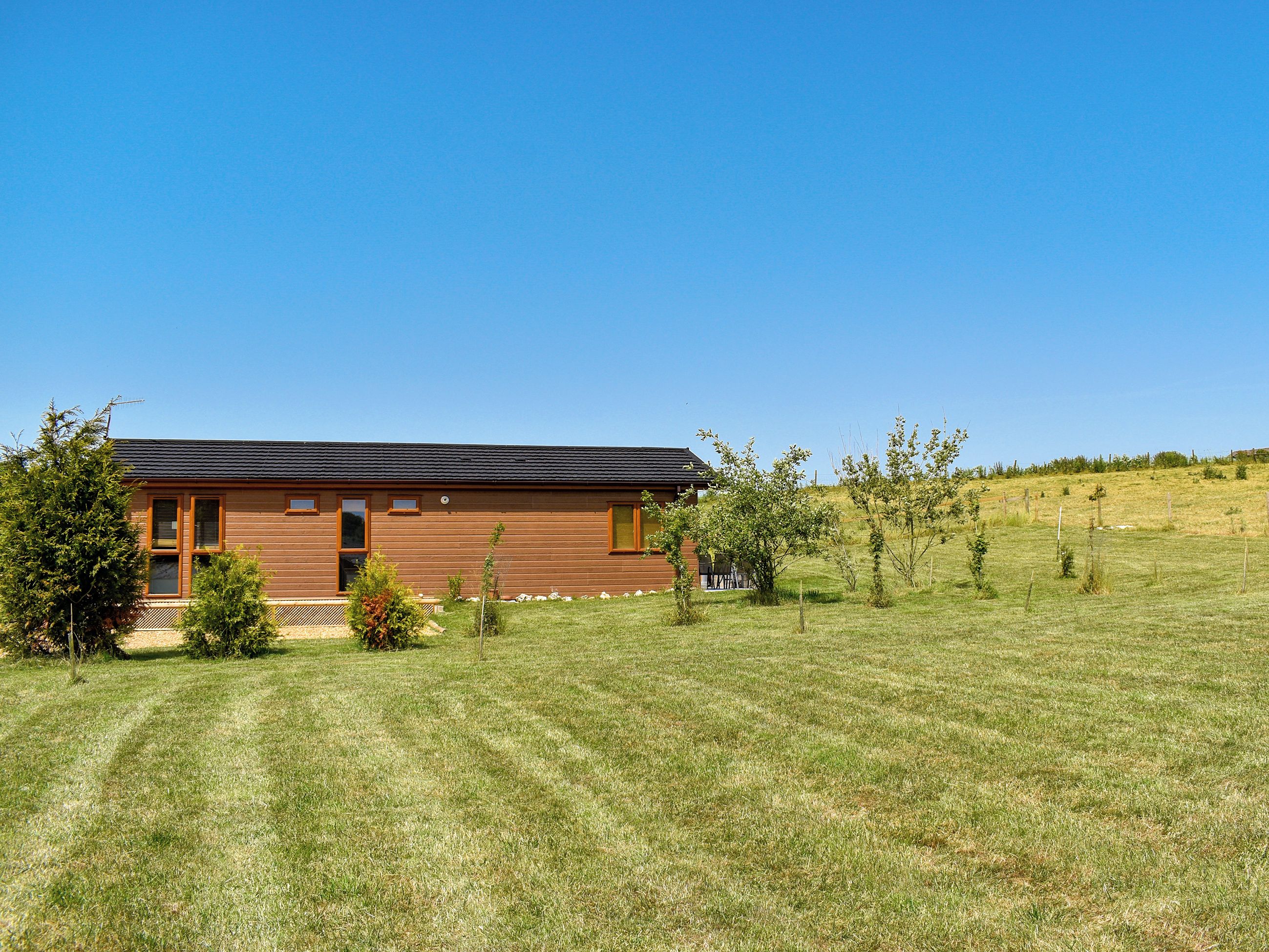 A single-story wooden house with a dark roof, surrounded by a grassy field and sparse trees.