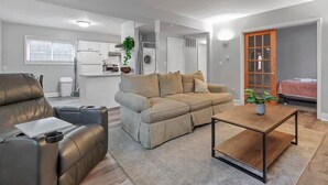 A modern living room with a sofa, a leather chair, and a coffee table. There is a kitchen area with appliances and a plant. - LowerFlat in GermanVillage - 3BR Pet | Yard | Park (Columbus)