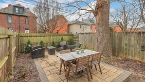 A backyard patio with a wooden table and chairs, surrounded by a wooden fence and a large tree. - LowerFlat in GermanVillage - 3BR Pet | Yard | Park (Columbus)