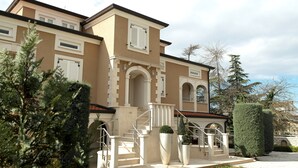 Un bâtiment à deux étages, doté d’une façade beige, de bordures blanches et d’un toit rouge. On y trouve de nombreuses fenêtres, un escalier avec des rampes blanches et une pelouse bien entretenue, agrémentée d’arbres à feuilles persistantes.