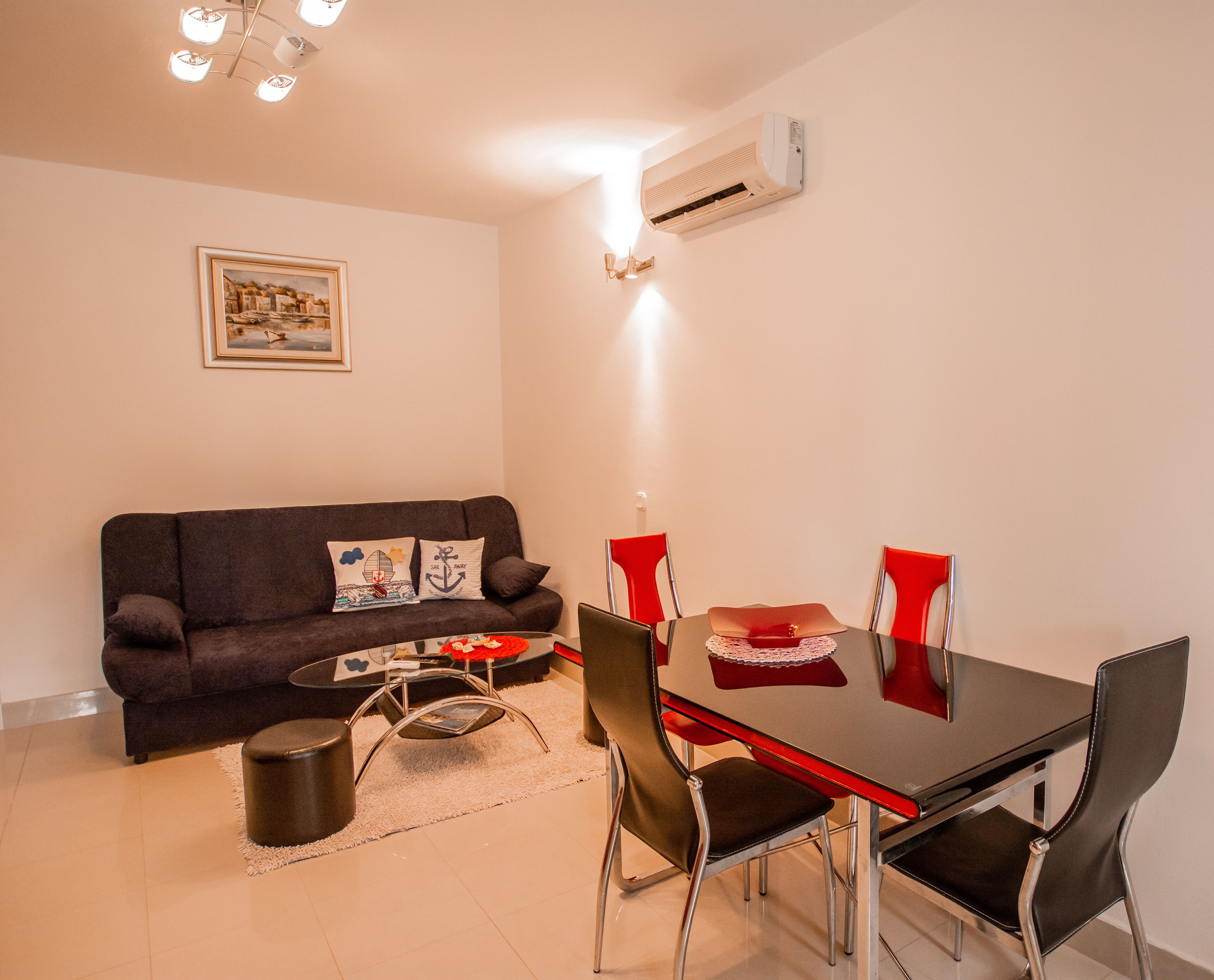 Uma sala de estar moderna com um sofá marrom escuro, uma mesa de jantar de vidro com cadeiras vermelhas, uma pequena mesa de centro e um ar condicionado branco fixado na parede.