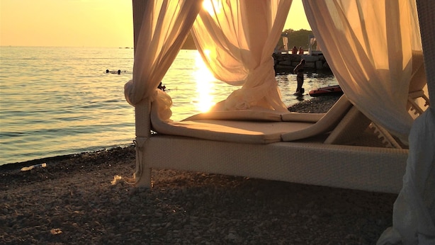Un lit installé sur la plage, agrémenté de rideaux légers, avec vue sur une mer calme et un coucher de soleil.