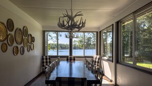 Un coin repas avec une grande table, des chaises à carreaux et un lustre. De grandes fenêtres offrent une vue sur un lac et des arbres.