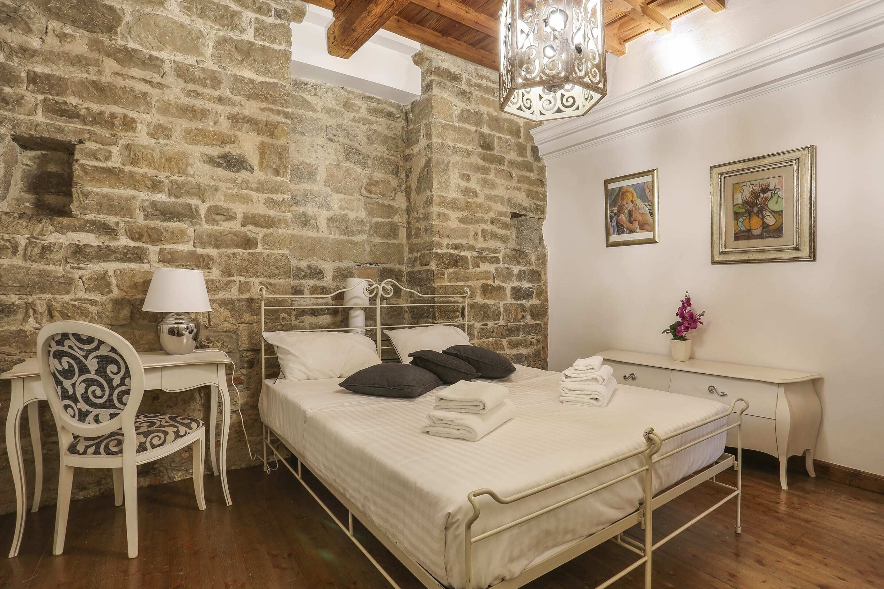 A bedroom with a stone wall, a metal bed frame, a white desk, a chair, and a hanging light fixture.