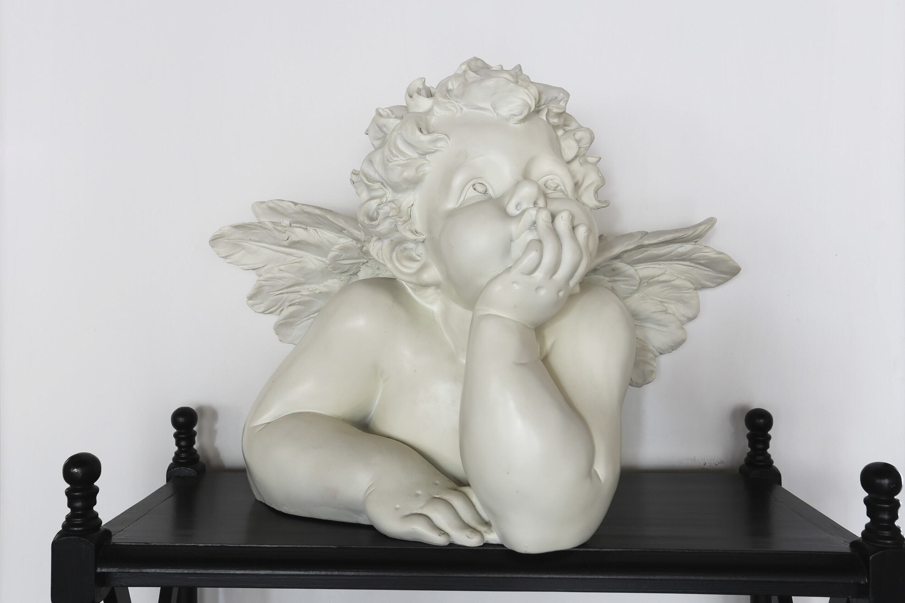 A white sculpture of a cherub with wings on a black table against a plain wall.