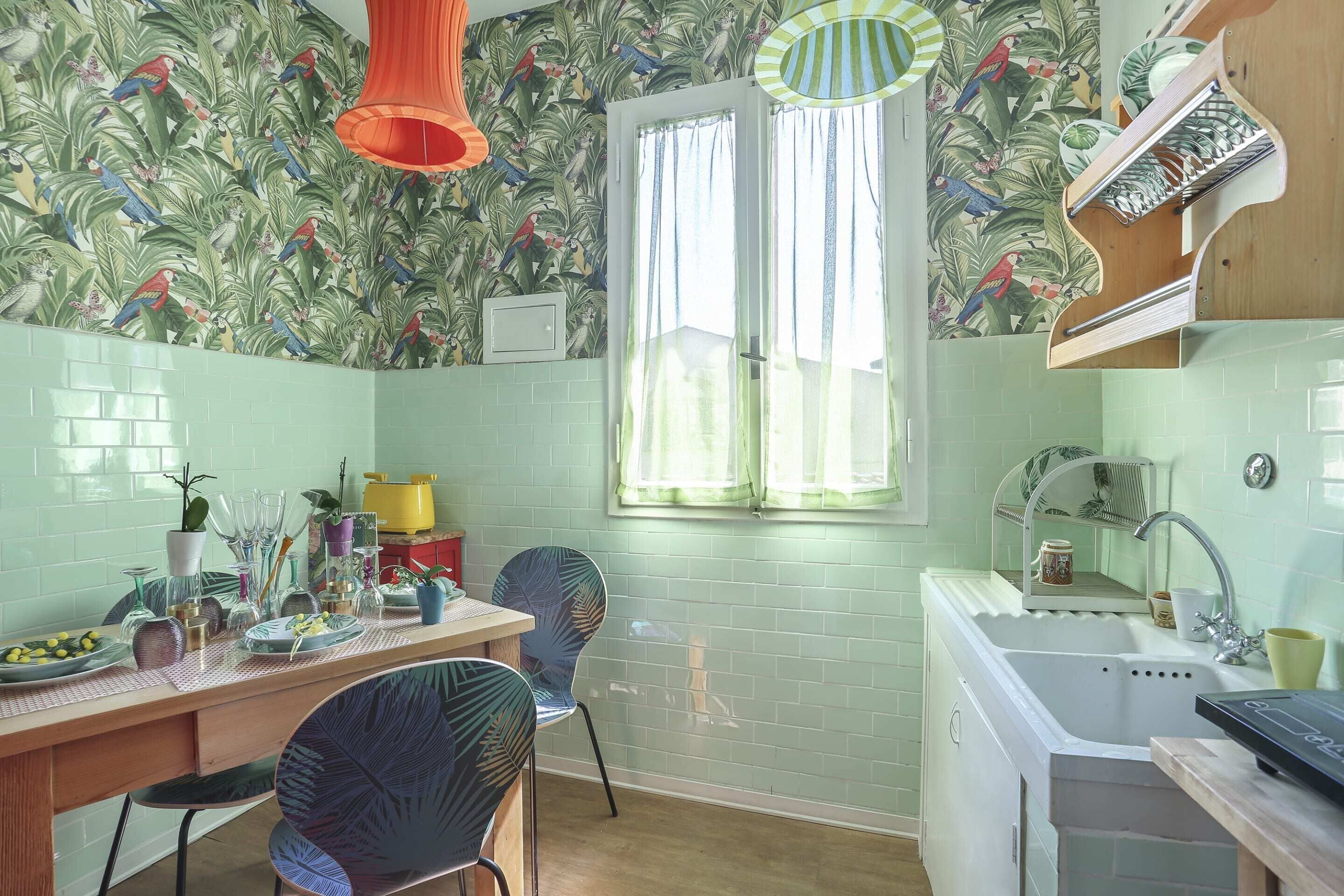 Una cocina con salpicadero de azulejos, una ventana con cortinas, un lavamanos y una zona de comedor con sillas y una mesa.