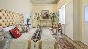 A bedroom with a tufted headboard, a bed with a decorative bedspread, a framed painting above the fireplace, and a view of the outside through an open door. - Lullaby Suite (Florence)