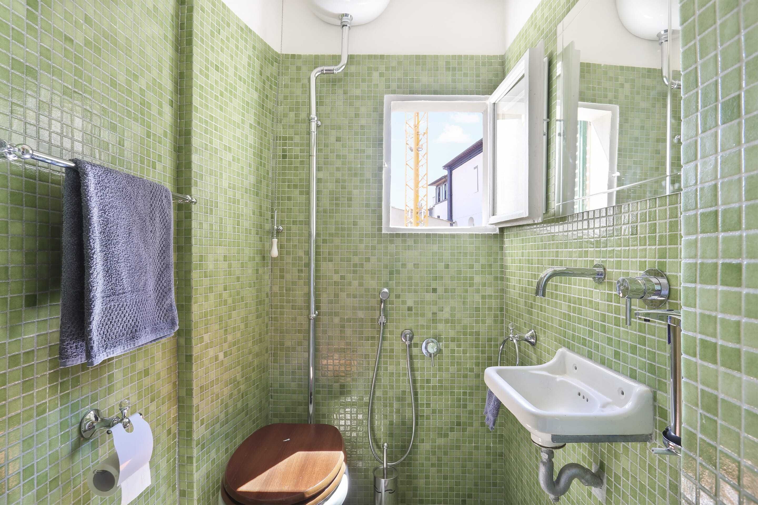 Une salle de bain avec des carreaux de mosaïque verts, des toilettes en bois, un lavabo blanc et une fenêtre donnant sur un immeuble.