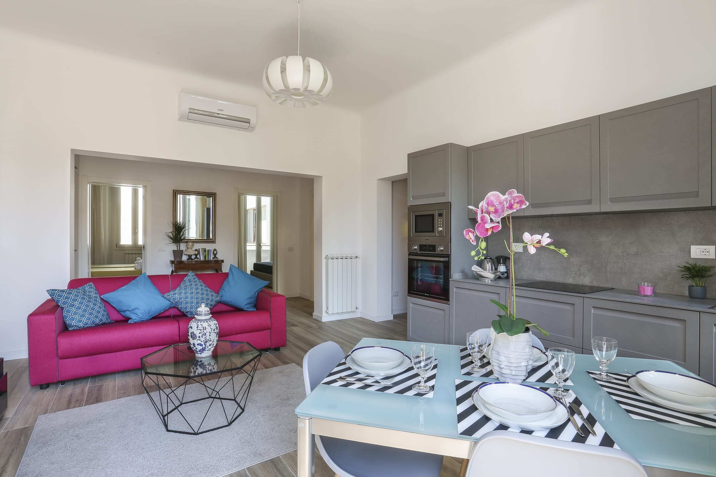 A modern living room with a pink sofa, a glass coffee table, and a dining area with striped placemats and white plates.