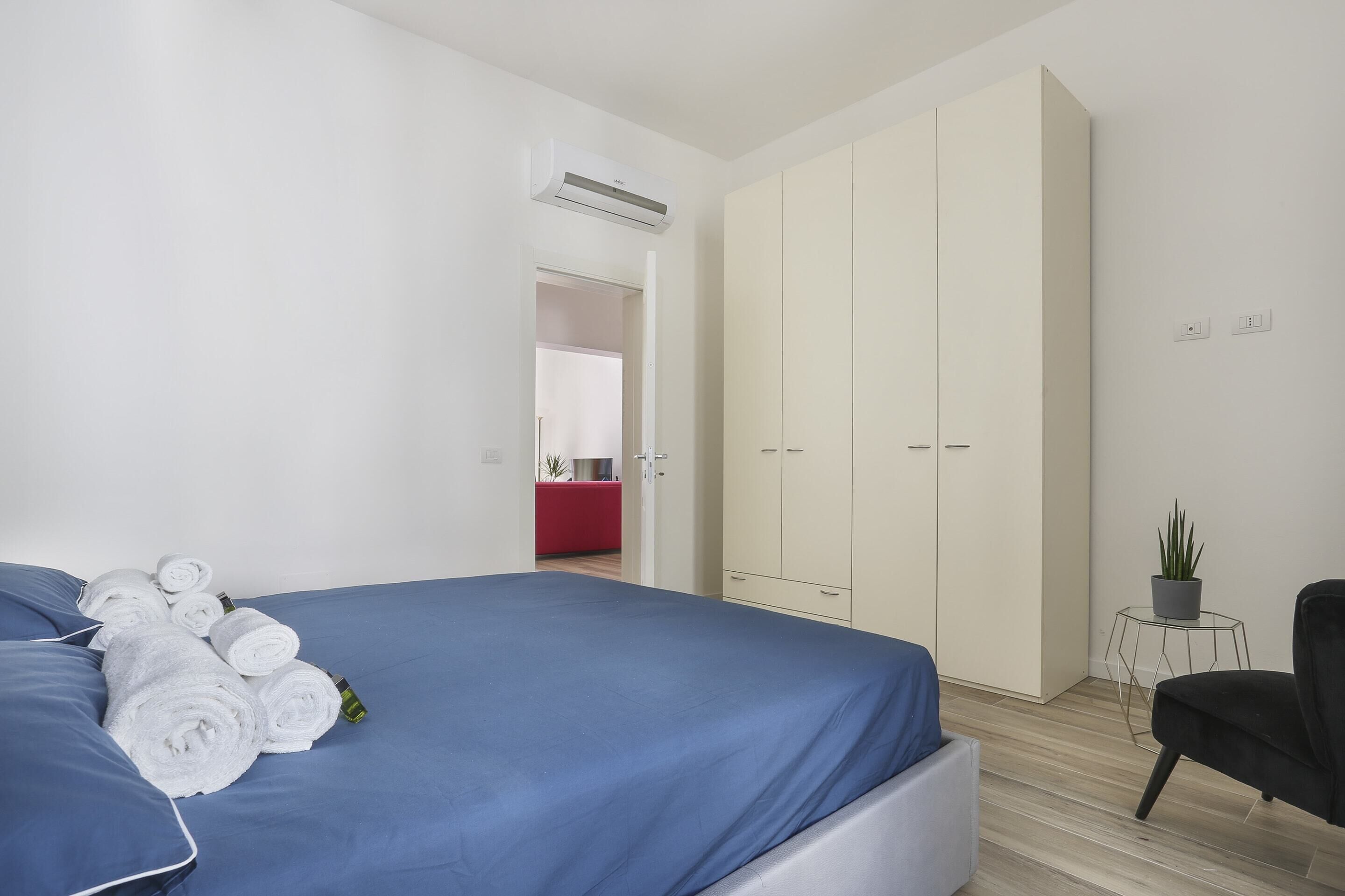 A modern bedroom with a large bed, a wardrobe, a small table with a plant, and a red piece of furniture in the background.