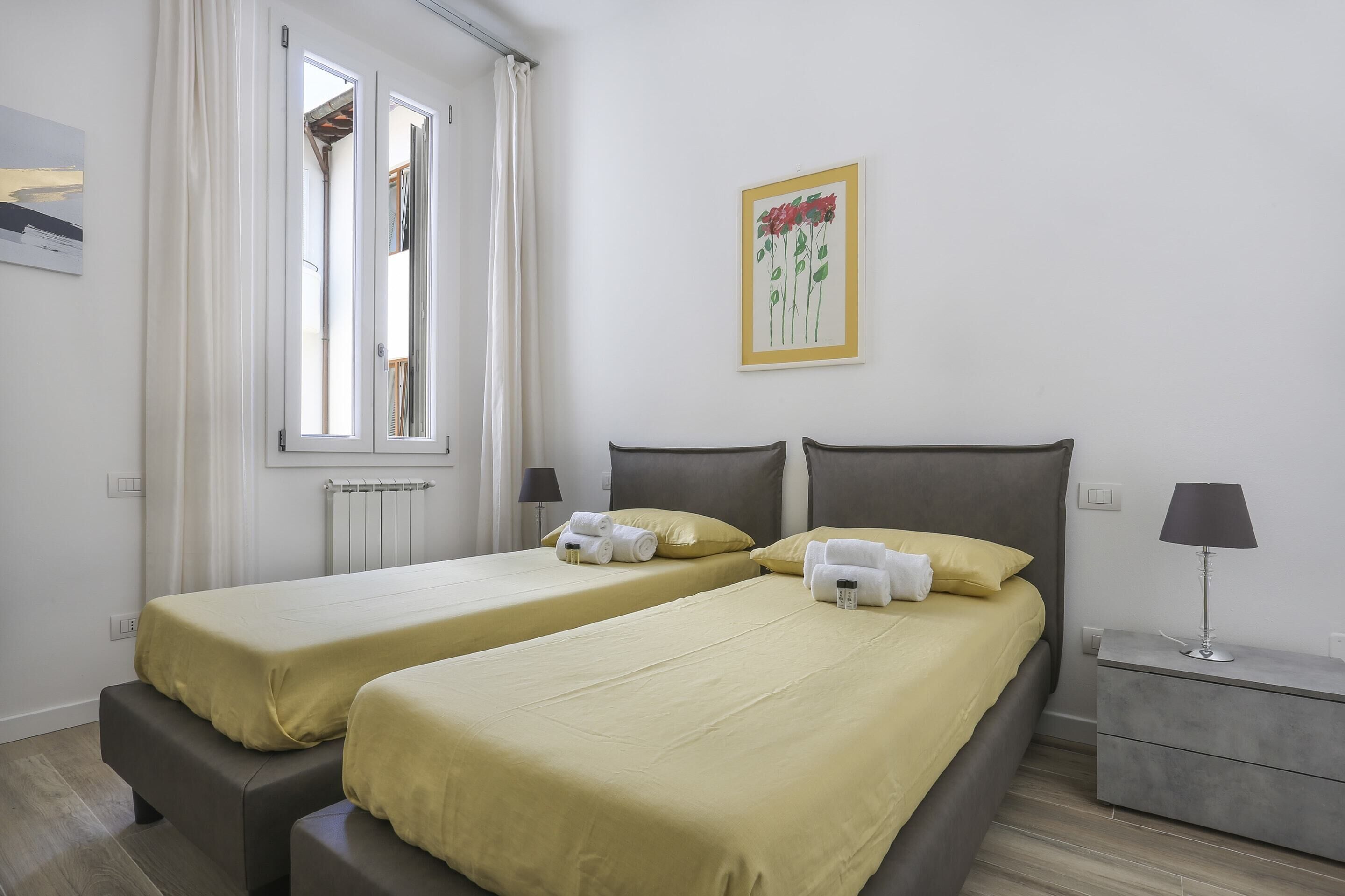 Una habitación de hotel con dos camas individuales, una mesita de noche con lámpara, una ventana con cortinas y un cuadro en la pared.
