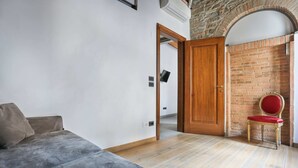 Una habitación con un sofá gris, un sillón tapizado de rojo y una puerta de madera que da a un muro de ladrillo.