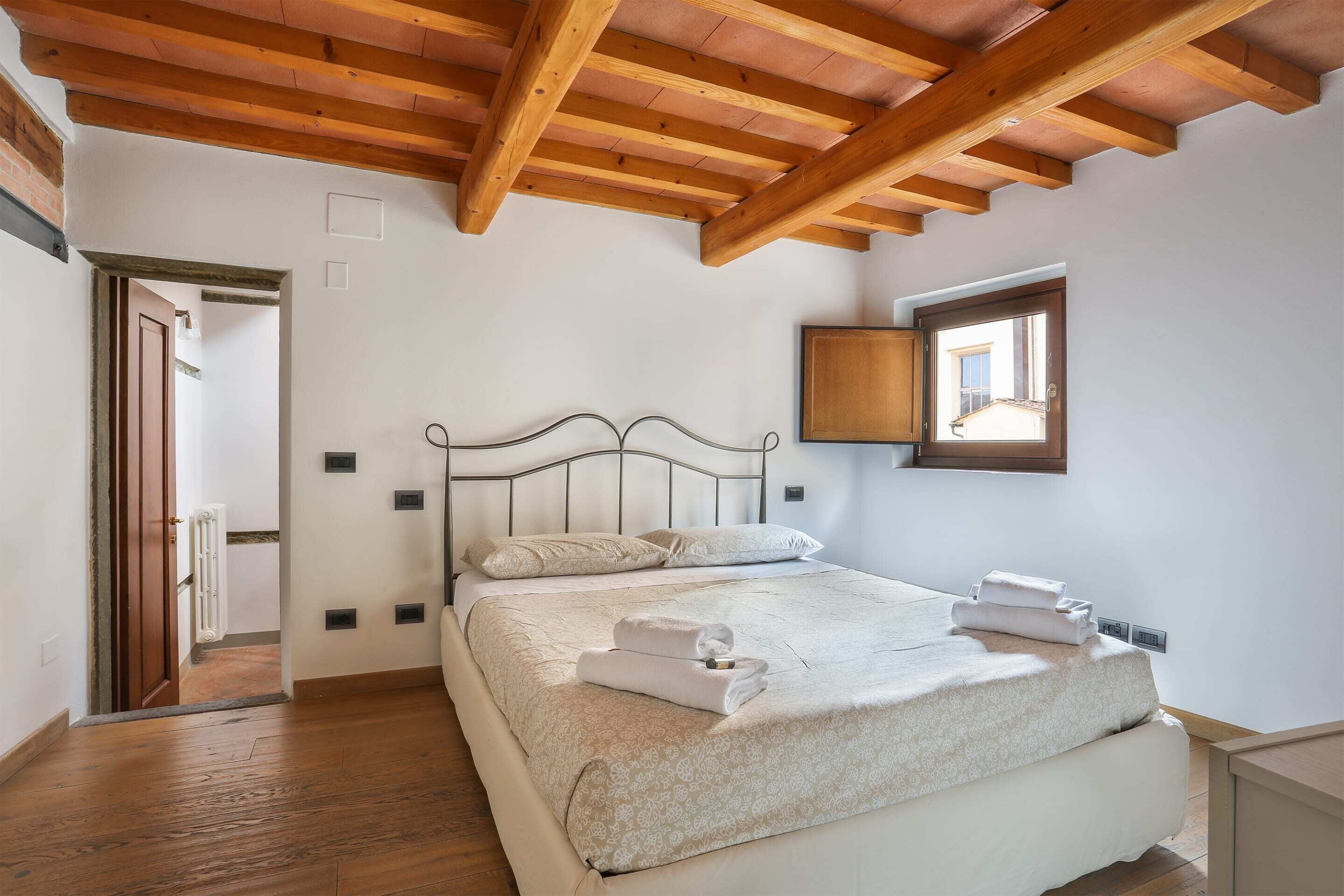 Une chambre avec un plafond en bois, un lit doté d’une tête de lit blanche et une fenêtre donnant sur un bâtiment à l’extérieur.