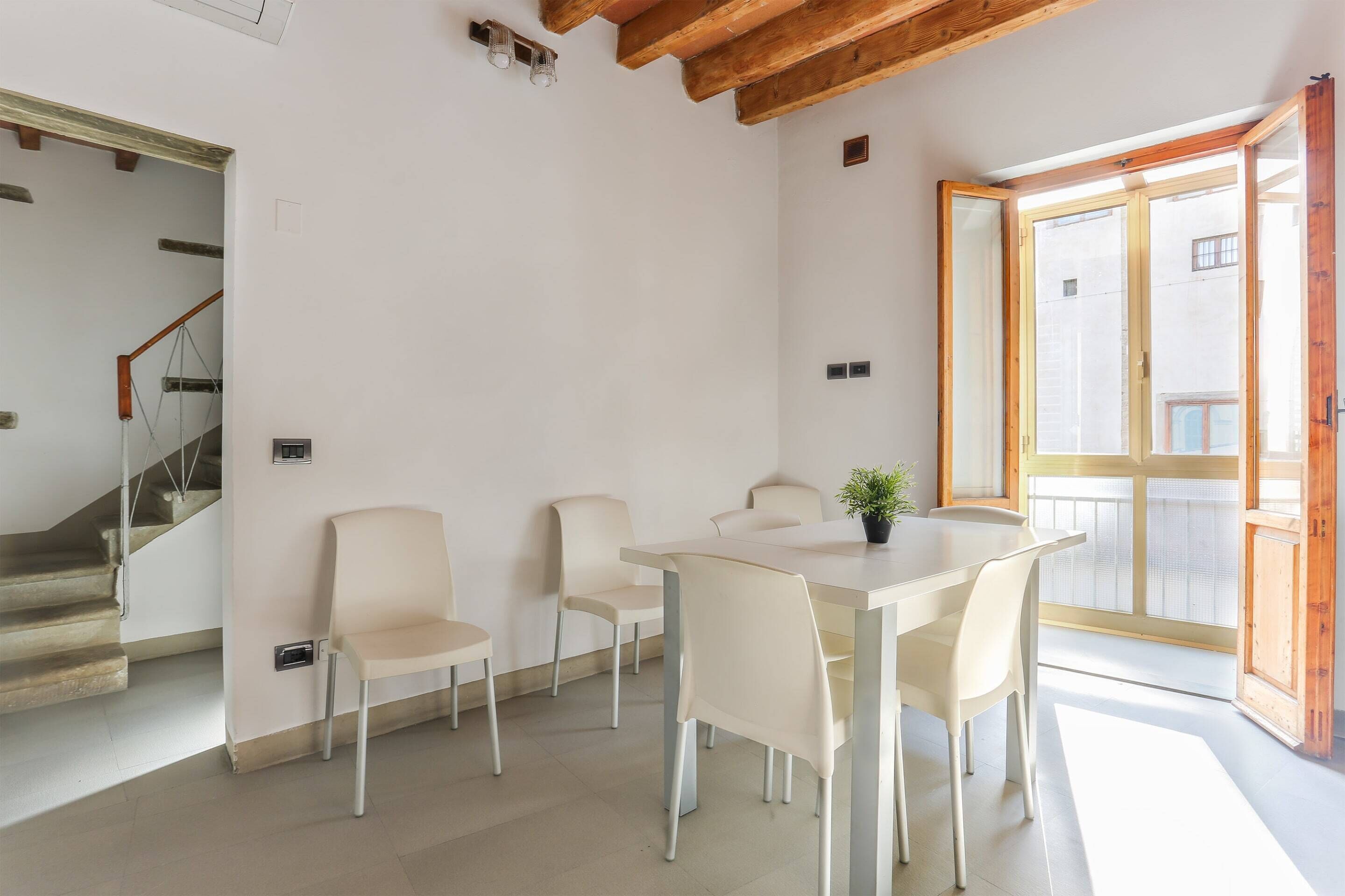 A modern dining area with a white table and chairs, a potted plant, and a staircase leading to an upper floor.