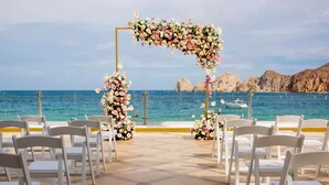 An outdoor wedding ceremony setup with white chairs and floral arches by the sea.