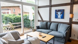 Uma sala de estar moderna com um sofá cinza, uma mesa de centro de madeira e uma janela grande com vista para uma casa de tijolos e vegetação.