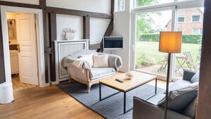Uma sala de estar aconchegante com um sofá, uma mesa de centro e uma televisão. Há uma janela com vista para um jardim e um edifício de tijolos.