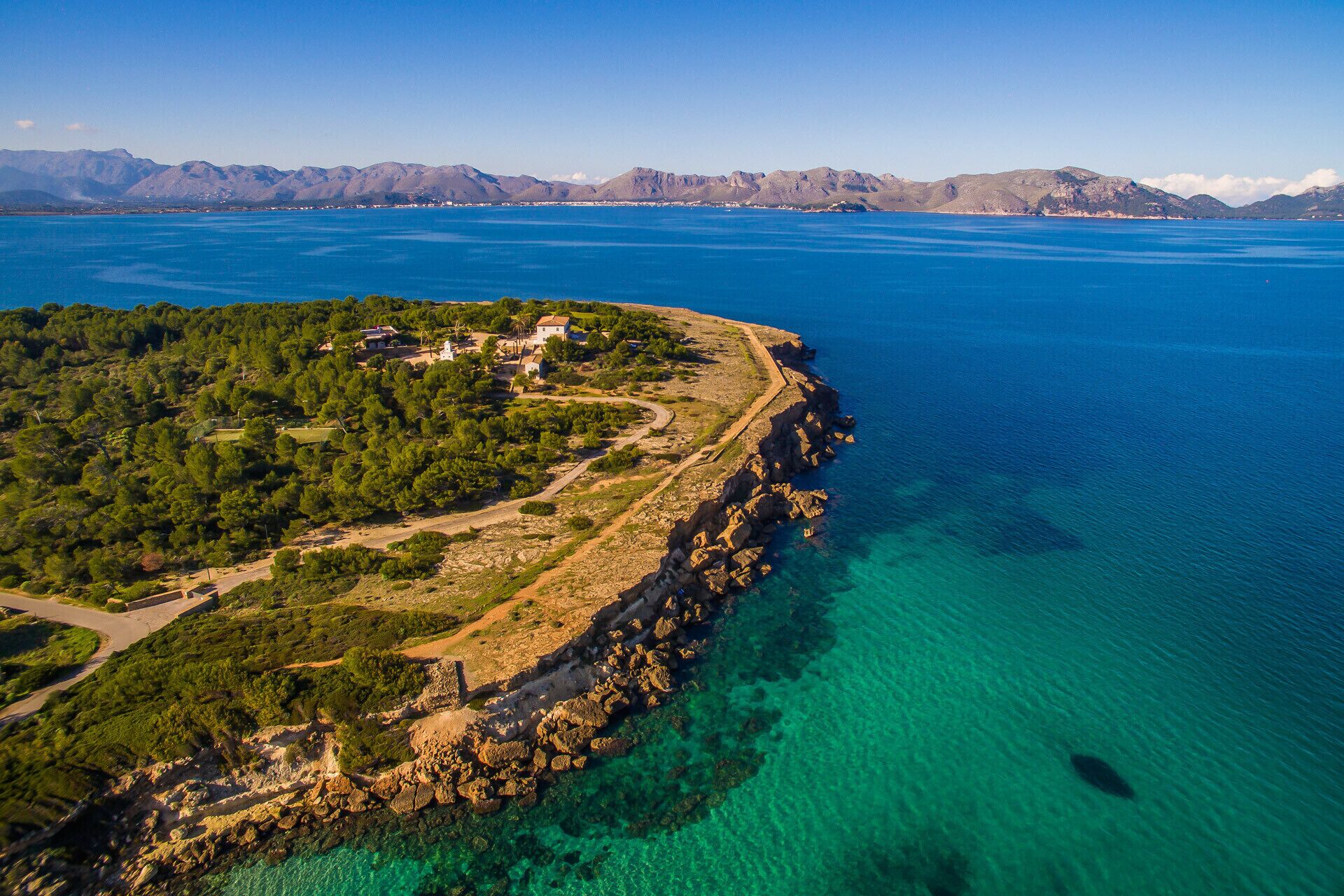 An aerial view of a coastal area with a small settlement, clear blue waters, and mountainous background.