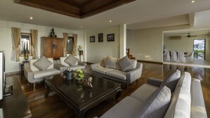 Ein geräumiges Wohnzimmer mit hoher Decke, Holzbalken und Deckenventilator. Zwei Sofas und ein Couchtisch mit Dekoration sind zu sehen. Außerdem ein Schrank und ein Fernseher.