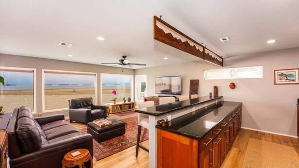 A spacious living area with a large window overlooking a beach, a dark leather sofa, a wooden coffee table, and a kitchen area with a black countertop and wooden cabinets. - Casa Azul (Seal Beach)