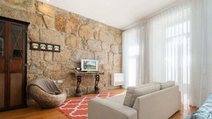Un salon avec un mur en pierre, un canapé, un fauteuil en osier, une petite table sur laquelle est posée une photo, et un tapis rouge.