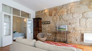 Un salon avec un mur en pierre, un canapé, un fauteuil en osier, un téléviseur posé sur un meuble en bois et une porte donnant sur une autre pièce.