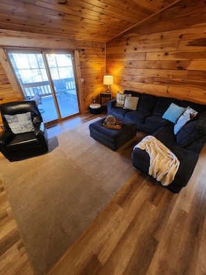 A cozy cabin interior with a wooden ceiling, a black leather chair, a dark-colored sofa, and a small table with a lamp.