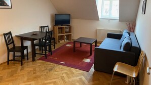Ein Wohnzimmer mit Holzdielen im Fischgrätmuster, einem dunklen Sofa, einem kleinen Tisch, einem Fernseher auf einem Holzschrank und einem Fenster mit Blick auf Gebäude.