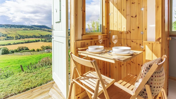 A wooden cabin with a dining table set for two, overlooking a green landscape. - Woodside Hut 1 - UK36105 (Strathpeffer)