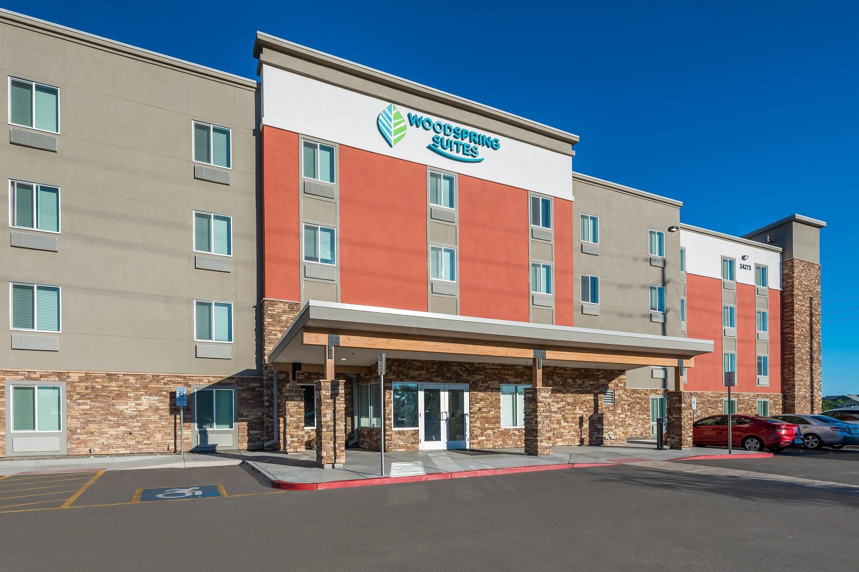 Un hotel de dues plantes amb façana moderna, un cartell ben visible de Woodspring Suites i un aparcament amb espais delimitats.