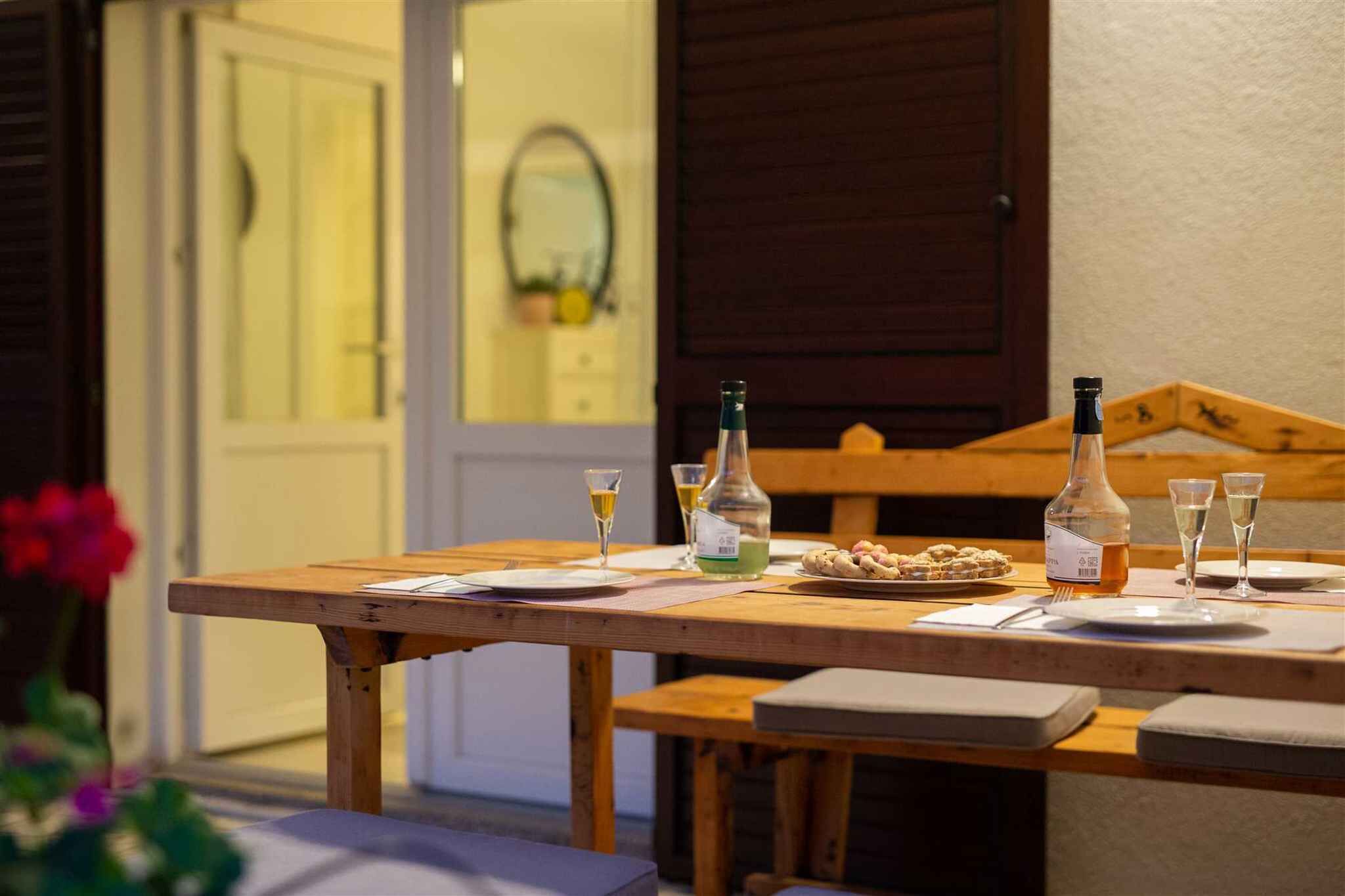 A wooden table set with plates, glasses, and bottles, with a bench and cushions in the foreground.