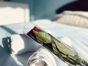A neatly made bed with a rolled white towel and a bouquet of flowers, including a red rose, on a white pillow.